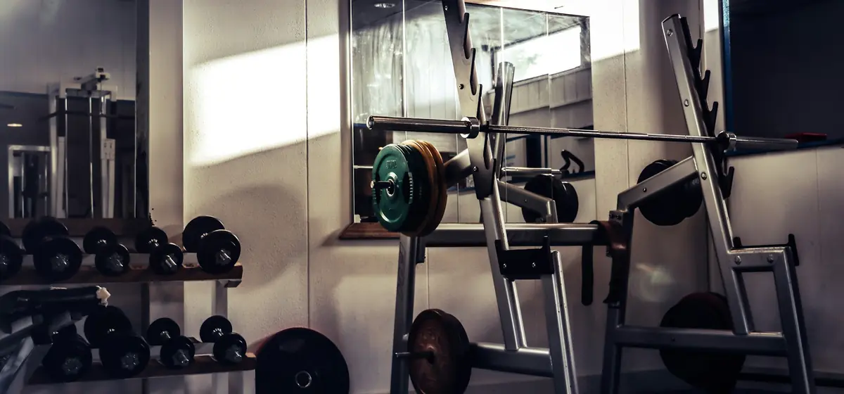 Rack de agachamento com barra, anilhas e halteres ao fundo em uma sala de treino